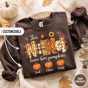 May include: A dark chocolate brown crewneck sweatshirt featuring the text "This NANA loves her pumpkins" in white script. The word "NANA" is decorated with autumn patterns, including plaid, polka dots, and leaves. Three orange pumpkins are printed below with the names "Josh", "Olivia", and "Matt".