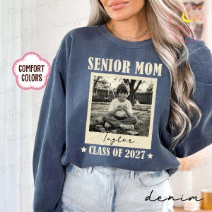 Może przedstawiać: Dżinsowa niebieska bluza z napisem "SENIOR MOM" nad czarno-białym zdjęciem dziecka. Zdjęcie jest podpisane "Taylor" i "CLASS OF 2027". Bluza ma r&oacute;wnież r&oacute;żowo-białą etykietę z napisem "COMFORT COLORS".