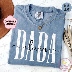 Könnte beinhalten: Ein blaues Jeans-T-Shirt mit dem Wort "DADA" in weißen Buchstaben und dem Namen "olivia" in schwarzer Schrift darunter. Das Shirt ist von Comfort Colors.