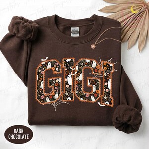 K&ouml;nnte beinhalten: Ein dunkel schokoladenbraunes Sweatshirt mit Rundhalsausschnitt und dem Schriftzug GIGI in gro&szlig;en Blockbuchstaben. Die Buchstaben sind mit einem Muster aus wei&szlig;en Geistern, orangefarbenen und rosa Blumen sowie Laub auf schwarzem Grund gef&uuml;llt.