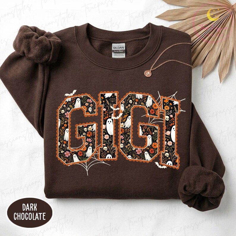 K&ouml;nnte beinhalten: Ein dunkel schokoladenbraunes Sweatshirt mit Rundhalsausschnitt und dem Schriftzug GIGI in gro&szlig;en Blockbuchstaben. Die Buchstaben sind mit einem Muster aus wei&szlig;en Geistern, orangefarbenen und rosa Blumen sowie Laub auf schwarzem Grund gef&uuml;llt.