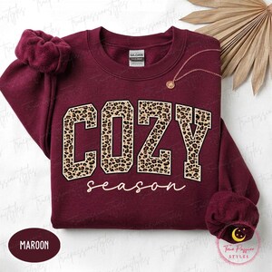 May include: A maroon long-sleeve sweatshirt featuring the word COZY in large block letters with a leopard print pattern and the word season in white cursive script below. The garment has a crew neckline and ribbed cuffs on the sleeves.