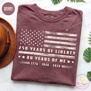 May include: Berry-coloured t-shirt with a distressed American flag design. The text reads "250 Years of Liberty" and "80 Years of Me," with the years 1776-1946-2026. The shirt has a crew neck and short sleeves.