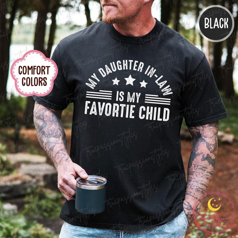 May include: Black t-shirt with the white text "MY DAUGHTER IN-LAW IS MY FAVORITE CHILD". The shirt has a crew neck and short sleeves. The text is arranged in an arc with three stars above the words "IS MY FAVORITE CHILD".