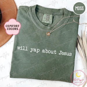 May include: A moss green Comfort Colors t-shirt with the text "will yap about Jesus" in white. A gold necklace with a pendant rests on the shirt. The shirt is folded and displayed with a hat and decorative elements.