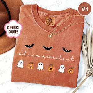 May include: A yam-colored Comfort Colors t-shirt with a crew neck and short sleeves. The shirt features black bat graphics above the words "admin assistant" and cartoon ghosts and pumpkins. A gold necklace is draped on the shirt.