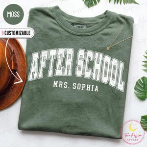 May include: A moss green t-shirt featuring the white distressed text AFTER SCHOOL in an arched collegiate font, with the name MRS. SOPHIA printed below in a smaller white sans-serif font. The shirt has a crew neckline and a short-sleeve design.