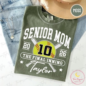 May include: A moss green t-shirt featuring white text that reads SENIOR MOM, 2026, and THE FINAL INNING, along with the name Taylor. A yellow softball graphic with the number 10 and two crossed baseball bats are printed in the center of the shirt.
