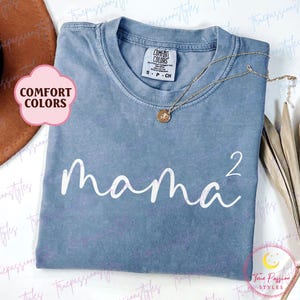 May include: A blue Comfort Colors t-shirt with the word "mama²" in white script. A gold necklace with a pendant is draped on the shirt. The shirt is folded neatly on a white surface.