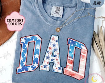 Pode incluir: Uma camiseta de jeans azul com a palavra "DAD" em letras brancas grandes. As letras são decoradas com um padrão de bandeira americana vermelho, branco e azul. A camiseta tem uma etiqueta que diz "Comfort Colors" e "S-P-CH".