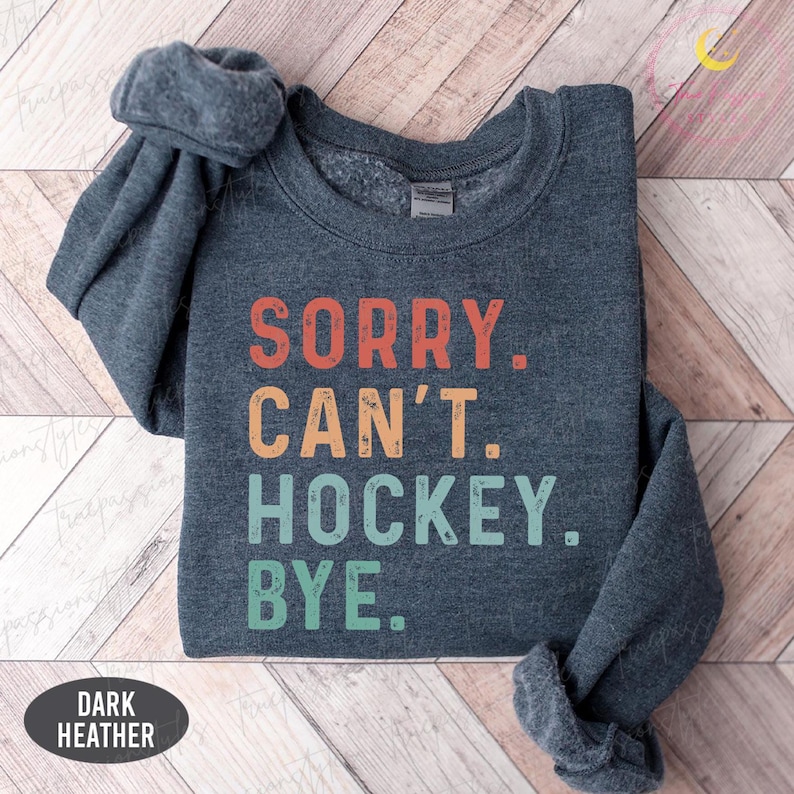 May include: Dark grey sweatshirt featuring the text "SORRY. CAN'T. HOCKEY. BYE." in orange, yellow, and green. The sweatshirt is folded, with the sleeves visible. The words "DARK HEATHER" are printed on a dark oval.