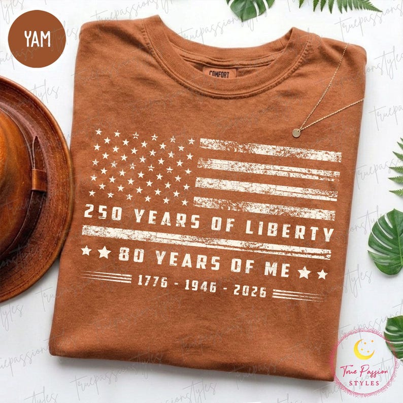 May include: A brown t-shirt with a distressed American flag graphic and the text "250 YEARS OF LIBERTY" and "80 YEARS OF ME", plus the years 1776-1946-2026. The shirt has a crew neck and a comfort tag.
