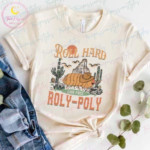 May include: A beige t-shirt with a graphic design of a cartoon roly-poly bug wearing a cowboy hat and the text "Roll Hard Live Free Roly-Poly".