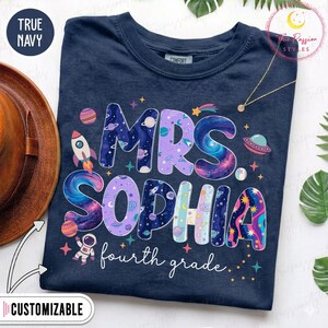 May include: A true navy short-sleeve t-shirt featuring the text MRS. SOPHIA and fourth grade in white script. The letters are filled with colorful space-themed graphics including planets, stars, a rocket ship, a UFO, and an astronaut.