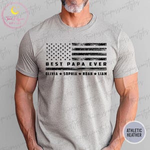 K&ouml;nnte beinhalten: Graues Athletic Heather T-Shirt mit einem verblassten amerikanischen Flaggen-Design in Schwarz. Unter der Flagge steht der Text "BEST PAPA EVER", darunter die Namen OLIVIA, SOPHIA, NOAH und LIAM. Das Shirt ist aus weichem Material.