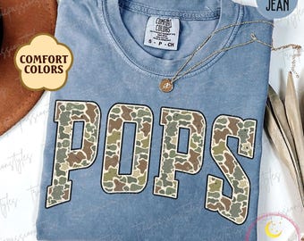 May include: A blue jean t-shirt with a camouflage pattern that spells out "POPS" in white outlined letters. The shirt is made by Comfort Colors.