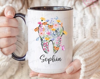 May include: White ceramic mug with a black interior and handle. The mug features a tooth-shaped design filled with colourful flowers and butterflies. The name "Sophia" is written in black script below the tooth design.