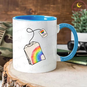 May include: White ceramic mug with a blue interior and handle. The mug features a colorful rainbow tea bag design with the text "LGBTQ" on the tag. The mug is sitting on a wooden surface.