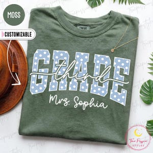 May include: A moss green t-shirt featuring the word GRADE in large, light blue block letters with white polka dots. The word third is written in white cursive script across the center, and Mrs. Sophia is printed in white below the text.