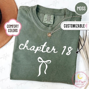 Peut inclure: Un t-shirt vert mousse avec les mots "chapter 18" et un nœud imprimés en blanc. Le t-shirt a un col rond et des manches courtes. Un collier en or avec un pendentif est sur le t-shirt. Les mots "Comfort Colors" sont sur une étiquette.