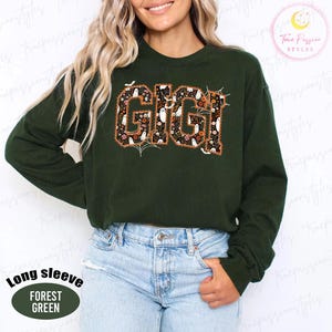 K&ouml;nnte beinhalten: Ein waldgr&uuml;nes Langarm-Sweatshirt mit dem Wort GIGI in gro&szlig;en Blockbuchstaben. Die Buchstaben sind mit einem Muster aus wei&szlig;en Geistern, orangefarbenen Blumen und Spinnweben auf schwarzem Grund gef&uuml;llt und haben eine d&uuml;nne orangefarbene Umrandung.