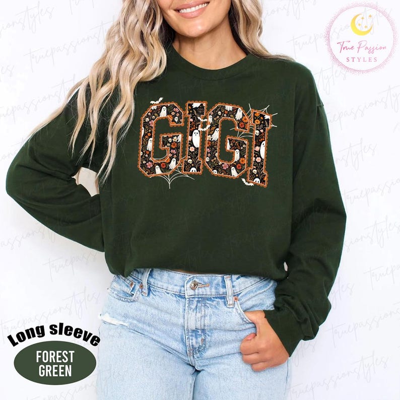 K&ouml;nnte beinhalten: Ein waldgr&uuml;nes Langarm-Sweatshirt mit dem Wort GIGI in gro&szlig;en Blockbuchstaben. Die Buchstaben sind mit einem Muster aus wei&szlig;en Geistern, orangefarbenen Blumen und Spinnweben auf schwarzem Grund gef&uuml;llt und haben eine d&uuml;nne orangefarbene Umrandung.