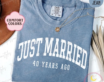 May include: Blue jean Comfort Colors t-shirt with the text "JUST MARRIED 40 YEARS AGO" in white. A gold necklace with a pendant is draped across the shirt. The shirt is folded and has a vintage look.