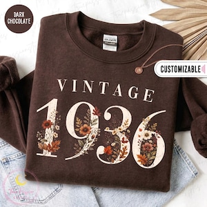 May include: A dark chocolate brown crewneck sweatshirt featuring the text VINTAGE 1936 in a cream-colored font. The numbers are filled with a detailed graphic of autumn-toned flowers and leaves in shades of orange, red, and brown.