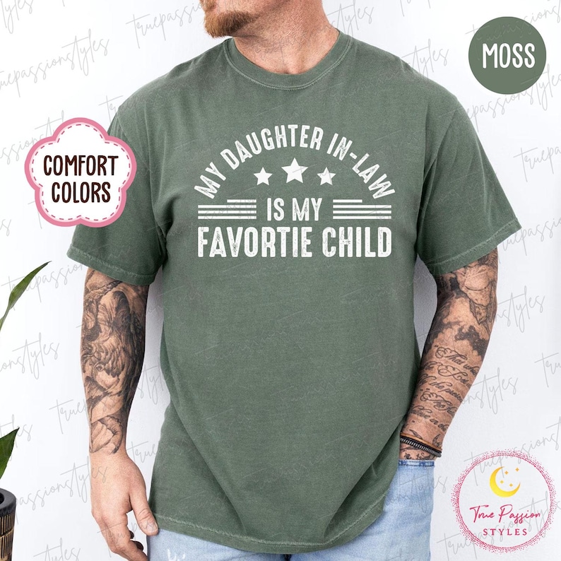 May include: A moss green t-shirt with the white text "MY DAUGHTER IN-LAW IS MY FAVORITE CHILD". The shirt has a crew neck and short sleeves. The words "COMFORT COLORS" are in a pink bubble on the left side of the shirt.