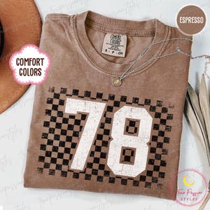 Könnte beinhalten: Espressofarbenes T-Shirt mit einem verblichenen Schachbrettmuster und der weißen Zahl 78. Das Shirt hat ein Comfort Colors-Etikett und eine silberne Halskette. Der Gesamtstil ist lässig und vintage.