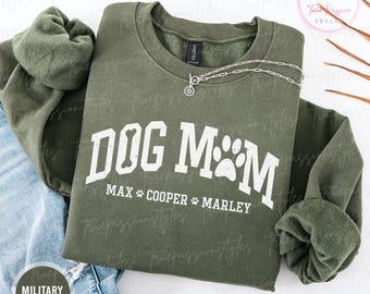 May include: Military green sweatshirt with the text "DOG MOM" in white block letters, a dog bone graphic, and paw prints. The names "MAX COOPER MARLEY" are printed below. A silver necklace is draped on the sweatshirt.