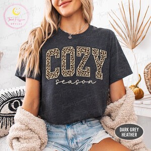 May include: A dark grey heather t-shirt featuring the word COZY in large block letters with a brown and black leopard print pattern, followed by the word season in a white cursive script font. The shirt has short sleeves and a crew neckline.