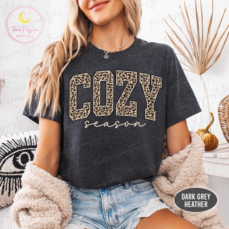 May include: A dark grey heather t-shirt featuring the word COZY in large block letters with a brown and black leopard print pattern, followed by the word season in a white cursive script font. The shirt has short sleeves and a crew neckline.