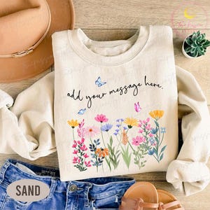 May include: Cream-colored sweatshirt with a floral design featuring pink, yellow, blue, and orange flowers, and butterflies. The text "add your message here" is written above the flowers. The sweatshirt is paired with denim shorts and sandals.