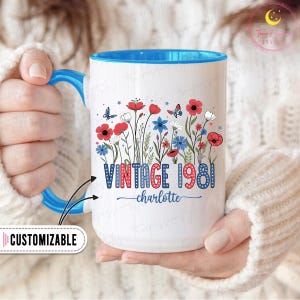May include: White ceramic mug with a blue interior and handle. The mug features a floral design with red, white, and blue flowers, butterflies, and the text "VINTAGE 1981" and "charlotte". The mug is customizable.
