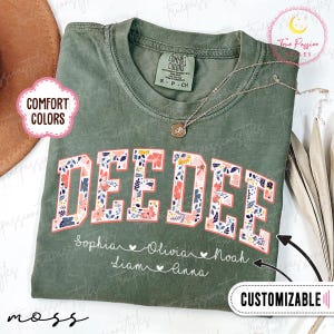 Könnte beinhalten: Ein moosgrünes T-Shirt mit dem Namen "DEEDEE" in einem floralen Design. Das Shirt enthält die Namen Sophia, Olivia, Noah, Liam und Anna. Eine goldene Halskette mit Anhänger ist ebenfalls sichtbar. Das Shirt ist anpassbar.