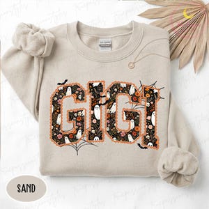 K&ouml;nnte beinhalten: Ein sandfarbenes Sweatshirt mit Rundhalsausschnitt und dem Schriftzug GIGI in gro&szlig;en Blockbuchstaben. Die Buchstaben sind mit einem Muster aus wei&szlig;en Geistern, orangefarbenen und rosa Blumen sowie schwarzem Laub gef&uuml;llt und orange umrandet.