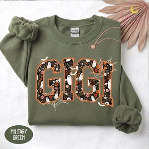 K&ouml;nnte beinhalten: Ein milit&auml;rgr&uuml;nes Sweatshirt mit Rundhalsausschnitt und dem Wort GIGI in gro&szlig;en Blockbuchstaben. Die Buchstaben sind mit einem dunklen Blumenmuster, wei&szlig;en Geistern und kleinen Totenk&ouml;pfen gef&uuml;llt. Das Design enth&auml;lt orangefarbene Umrisse, Spinnenweben und Flederm&auml;use.
