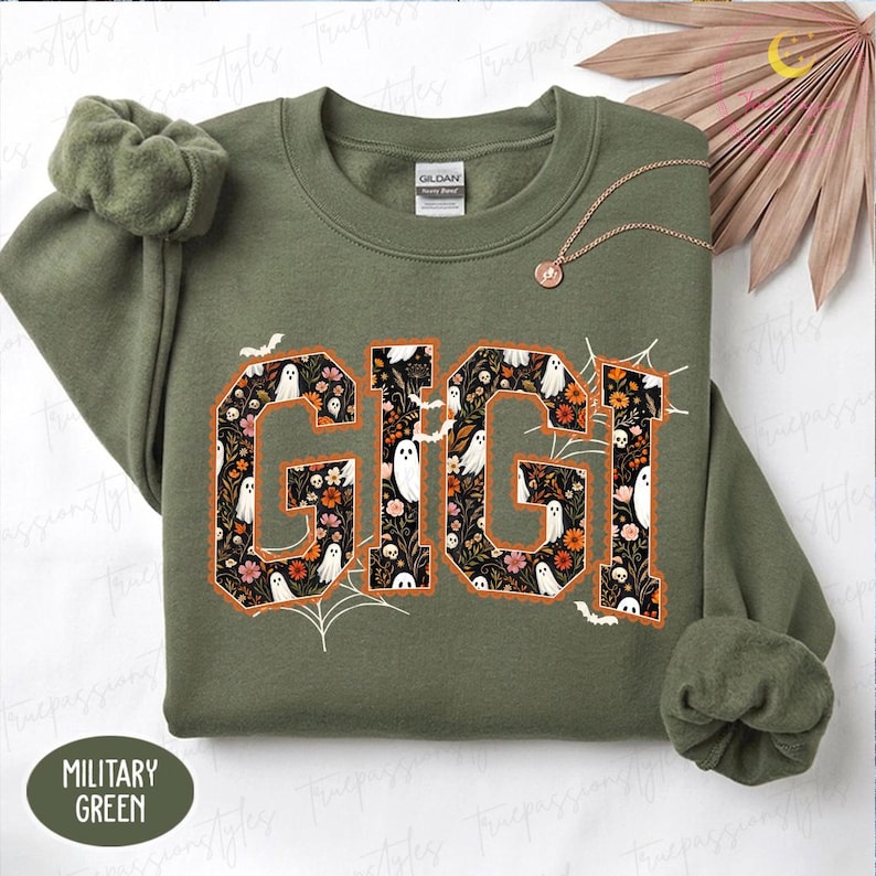K&ouml;nnte beinhalten: Ein milit&auml;rgr&uuml;nes Sweatshirt mit Rundhalsausschnitt und dem Wort GIGI in gro&szlig;en Blockbuchstaben. Die Buchstaben sind mit einem dunklen Blumenmuster, wei&szlig;en Geistern und kleinen Totenk&ouml;pfen gef&uuml;llt. Das Design enth&auml;lt orangefarbene Umrisse, Spinnenweben und Flederm&auml;use.