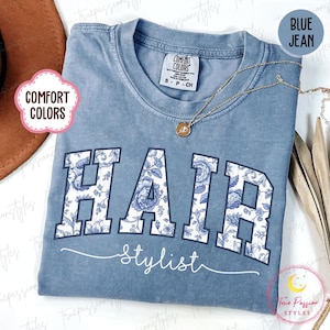 Puede incluir: Camiseta azul vaquero Comfort Colors con texto "HAIR stylist". La palabra "HAIR" tiene un diseño floral en negrita. Un collar dorado con un colgante está en la camisa. Talla pequeña.