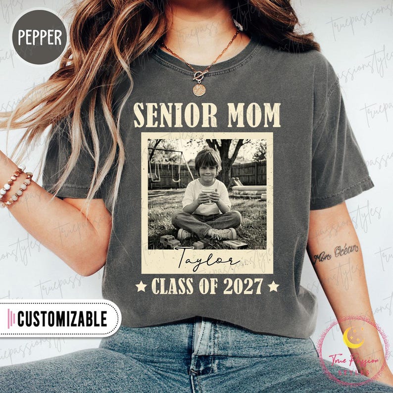 Może przedstawiać: Szara koszulka z napisem "SENIOR MOM" nad czarno-białym zdjęciem dziecka. Pod zdjęciem znajduje się imię "Taylor" i napis "CLASS OF 2027". Słowo "CUSTOMIZABLE" znajduje się na białej etykiecie.