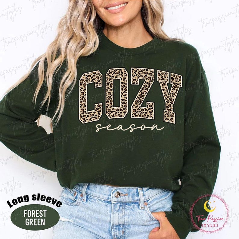 May include: A dark forest green long-sleeve sweatshirt featuring the word COZY in large, block-style letters with a brown and black leopard print pattern. Below the word COZY, the word season is written in a white, cursive script font.