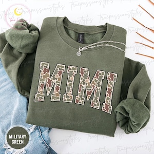 Könnte beinhalten: Ein olivgrünes Sweatshirt mit dem Wort "MIMI" in einem Camouflagemuster auf der Vorderseite. Eine silberne Halskette mit einem Sonnenanhänger liegt auf dem Sweatshirt. Die Worte "MILITARY GREEN" sind auf einem runden Etikett aufgedruckt.