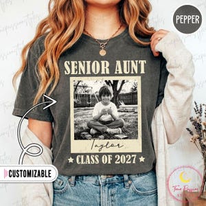 May include: A charcoal gray short-sleeve t-shirt featuring the text SENIOR AUNT in a vintage font above a black and white photograph of a child sitting on grass. Below the photo, the name Taylor and the text CLASS OF 2027 are printed in white.