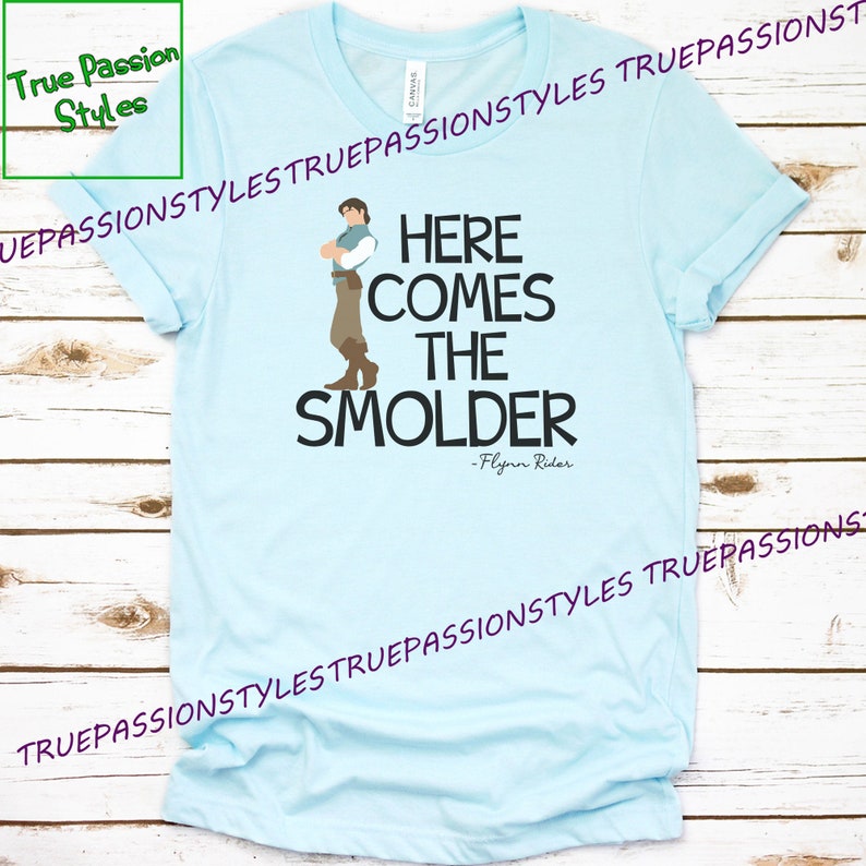 May include: Light blue t-shirt with a cartoon image of a man with brown hair and a brown outfit. The text "Here comes the Smolder - Flynn Rider" is printed on the shirt.