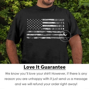 May include: Black t-shirt with a distressed white American flag design. The flag has the words "BRISKET BUTTS RIBS & FREEDOM" written on it. The text "Texas Passion Styles" is also on the shirt.