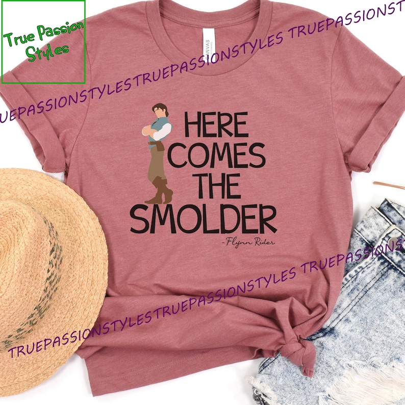 May include: A maroon t-shirt with a cartoon image of a man with brown hair and a blue shirt. The text "HERE COMES THE SMOLDER - Flynn Rider" is printed on the shirt.