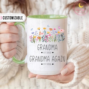 Op de afbeelding: Witte keramische mok met een groene handgreep en binnenkant. De mok heeft een bloemenontwerp met vlinders en de woorden "GRANDMA EST.2022" en "GRANDMA AGAIN EST.2026" in het zwart. Het woord "CUSTOMIZABLE" staat in een witte banner.
