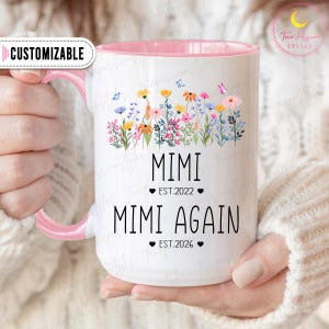 Könnte beinhalten: Weißer Keramikbecher mit rosa Innenraum und Henkel, verziert mit einem Blumendesign und Schmetterlingen. Der Becher hat den Text "MIMI" über "EST.2022" und "MIMI AGAIN" über "EST.2026". Das Wort "ANPASSBAR" steht auf dem Griff.