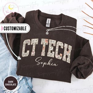 Könnte beinhalten: Dunkelbraunes Sweatshirt mit floralem "CT TECH"-Schriftzug und dem Namen "Sophia" in Schreibschrift. Das Sweatshirt hat einen Rundhalsausschnitt und lange Ärmel. Das Wort "Customizable" befindet sich in einem weißen Banner. Eine silberne Halskette befindet sich auf dem Sweatshirt.